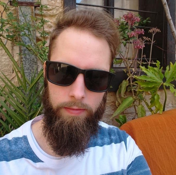 Ein Mann mit Sonnenbrille und Bart sitzt im Freien auf einer Terrasse.