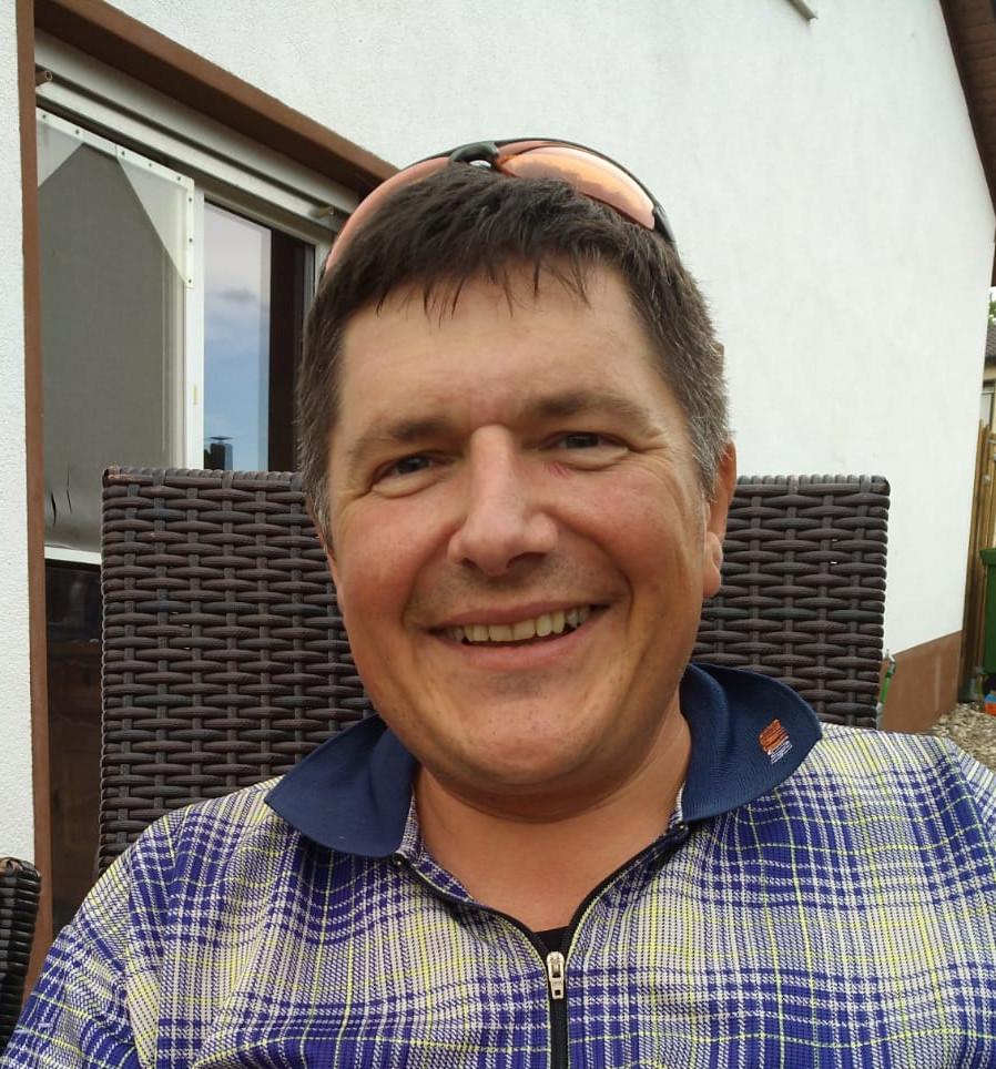 Ein lächelnder Mann mit kurz geschnittenen Haaren und einer Sonnenbrille auf dem Kopf. Er trägt ein blau-gelbes kariertes Hemd.