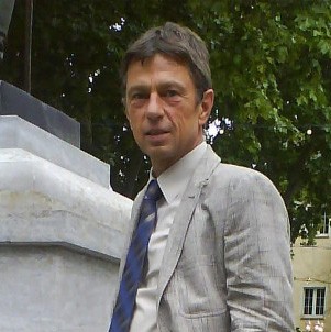 Ein Mann in einem hellen Anzug und dunkelblauer Krawatte steht neben einem Denkmal. Er hat kurzes braunes Haar.