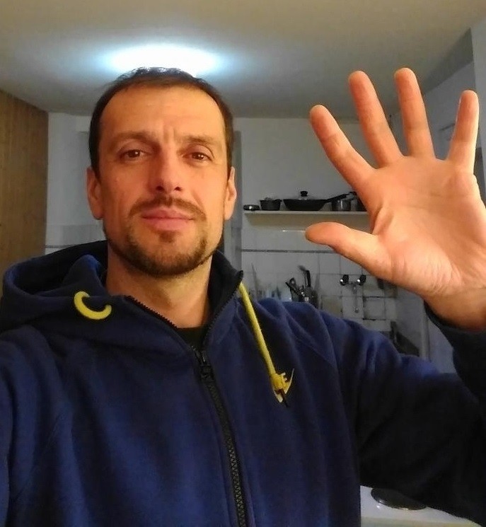 Ein Mann mit kurzem Haar und einem blauen Pullover mit gelben Details winkt mit seiner rechten Hand.