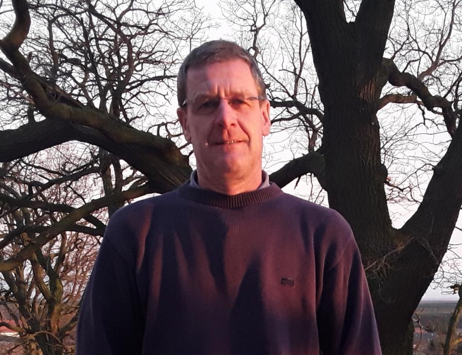 Ein Mann steht vor einem großen, kahlen Baum. Er trägt einen dunklen Pullover.