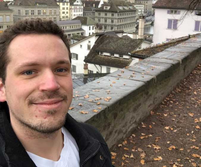 Ein Mann macht ein Selfie vor einer Stadt mit alten Häusern und einem Fluss. Der Mann trägt eine schwarze Jacke und ein weißes T-Shirt.