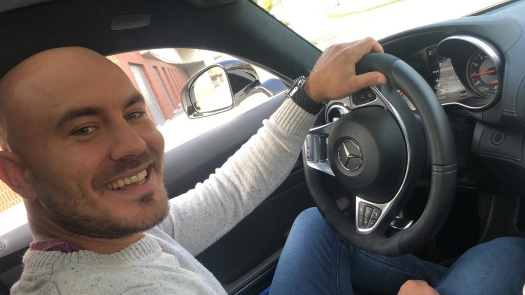 Ein Mann sitzt im Auto und lächelt. Er hält das Lenkrad in der Hand.