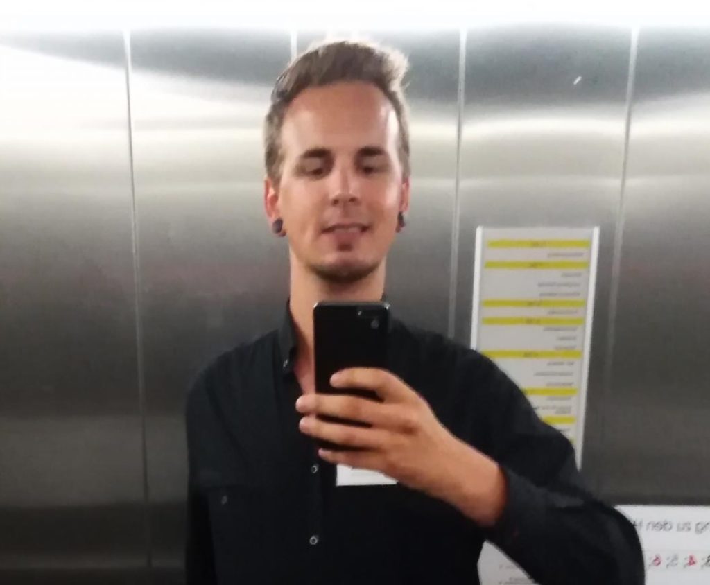 Ein Mann in einem schwarzen Hemd hält sein Telefon hoch, um ein Selfie im Aufzug zu machen. Er hat kurzes, braunes Haar und steht vor einer metallenen Wand.