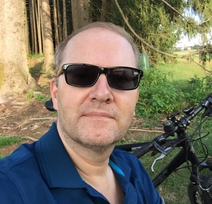 Ein Mann in einer blauen Polo-Shirt und Sonnenbrille steht neben einem Fahrrad im Freien.