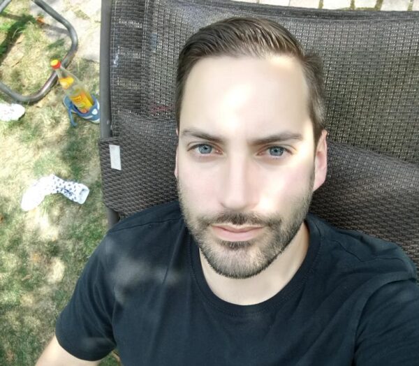 Ein Mann mit blauen Augen und einem gepflegten Bart sitzt auf einer Outdoor-Möbel. Er trägt ein schwarzes T-Shirt.