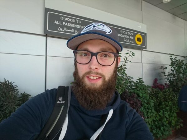 Ein Mann mit einem Bart und einer Kappe steht vor einem Flughafenschild.