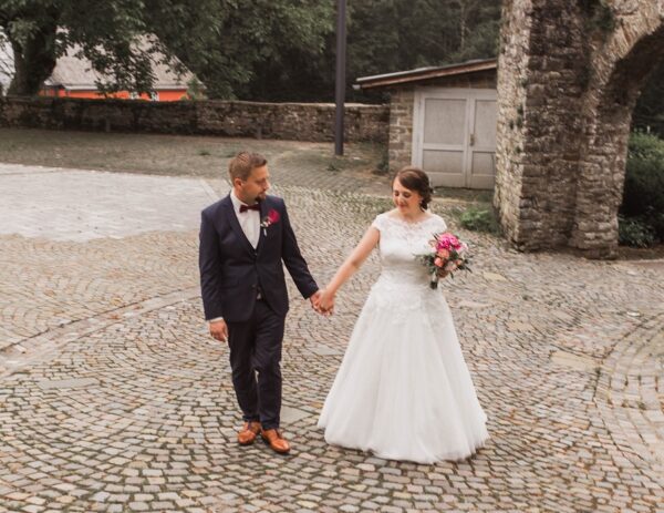 Ein frisch vermähltes Paar geht händchenhaltend über eine Kopfsteinpflasterstraße. Die Braut trägt ein weißes Kleid und einen Blumenstrauß.