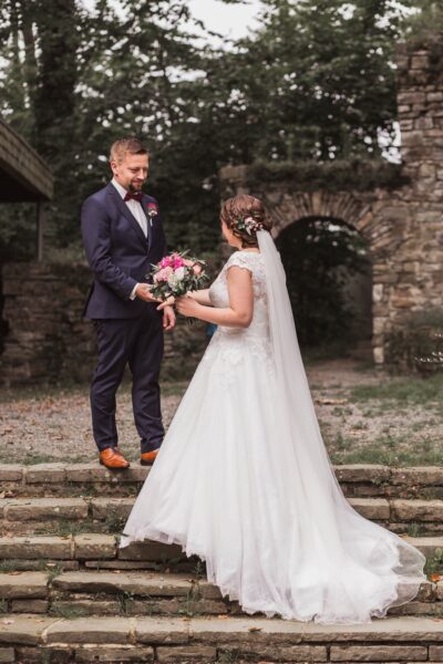 Ein Paar in Hochzeitskleidung, die Braut in einem weißen Kleid mit Blumensträußchen, der Bräutigam in einem dunklen Anzug.