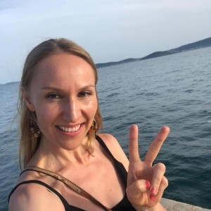 Eine lächelnde Frau macht einen Friedensgruß am Seeufer. Sie trägt ein schwarzes Sommerkleid und hat die Haare offen.