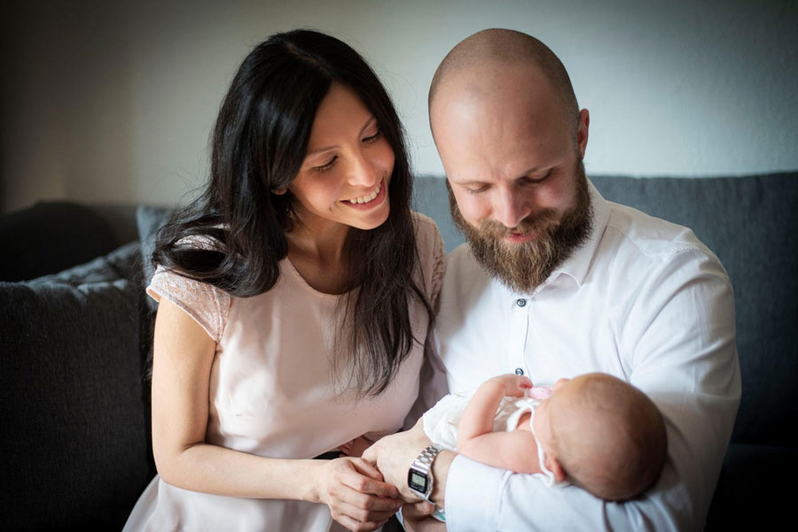 Ein Paar sitzt auf einem Sofa und hält ihr Baby. Die Mutter lächelt, während der Vater liebevoll auf das Kind schaut.